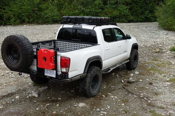 White off-road pickup truck equipped with roof rack and rear-mounted spare tire and red fuel container using BPF Universal Pelican Vault V800 and V730 Case Mounting brackets in rugged outdoor setting.