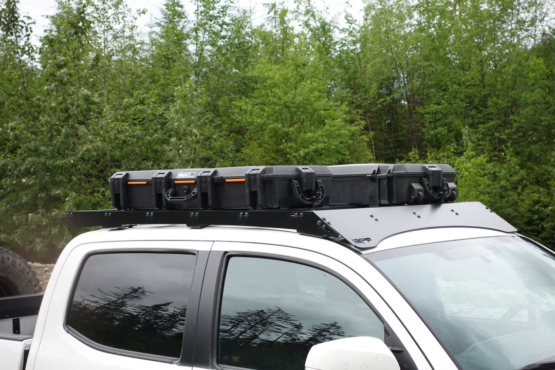 BPF Universal Pelican Vault V700 Case Mounting brackets securing a black Pelican Vault case on a white truck roof rack against a green forest background.