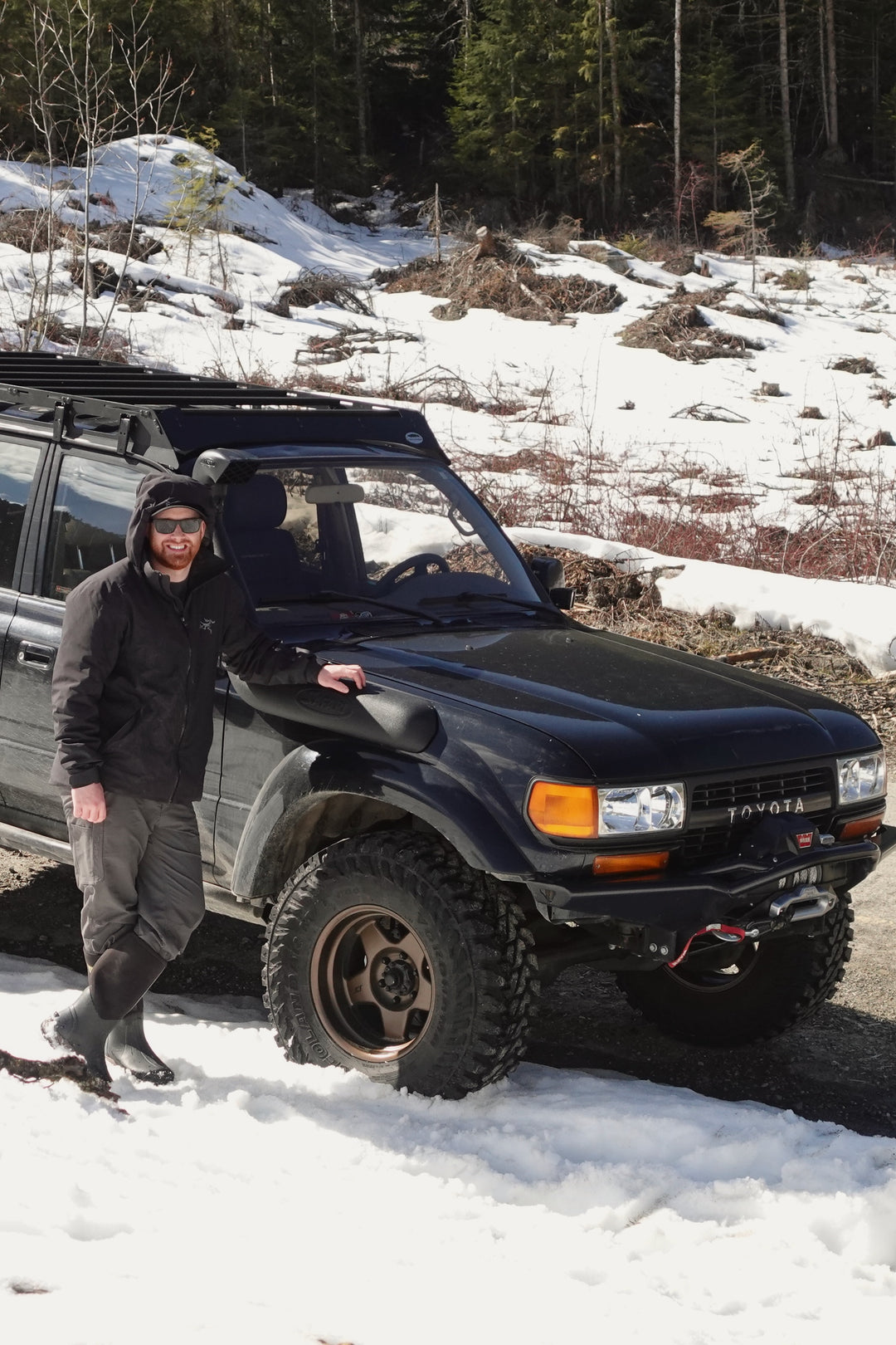 Man wearing outdoor gear and sunglasses standing next to a black Toyota SUV with off-road tires, parked on a snowy path with forest and snow-covered ground in the background.