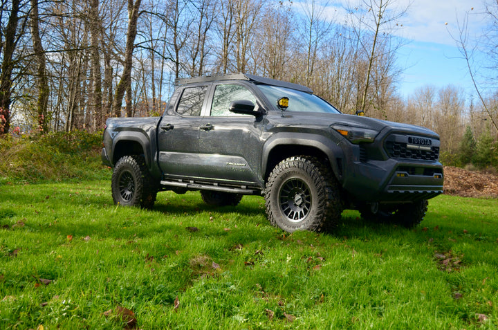 Black off-road pickup truck parked on grass with rugged tires and a 2024 Trailhunter Grille with BPF letters installed, surrounded by trees under a blue sky.