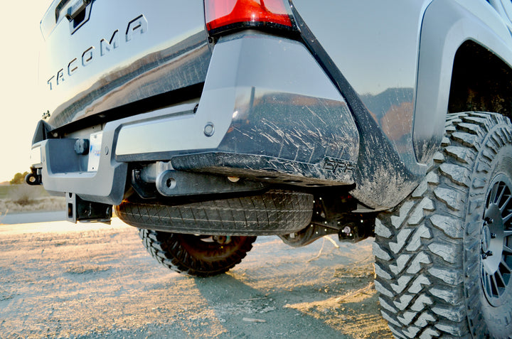 BPF 4th gen Tacoma rear tow hooks with integrated skid plates installed on a gray Tacoma truck showing heavy-duty off-road tires and rugged terrain background.