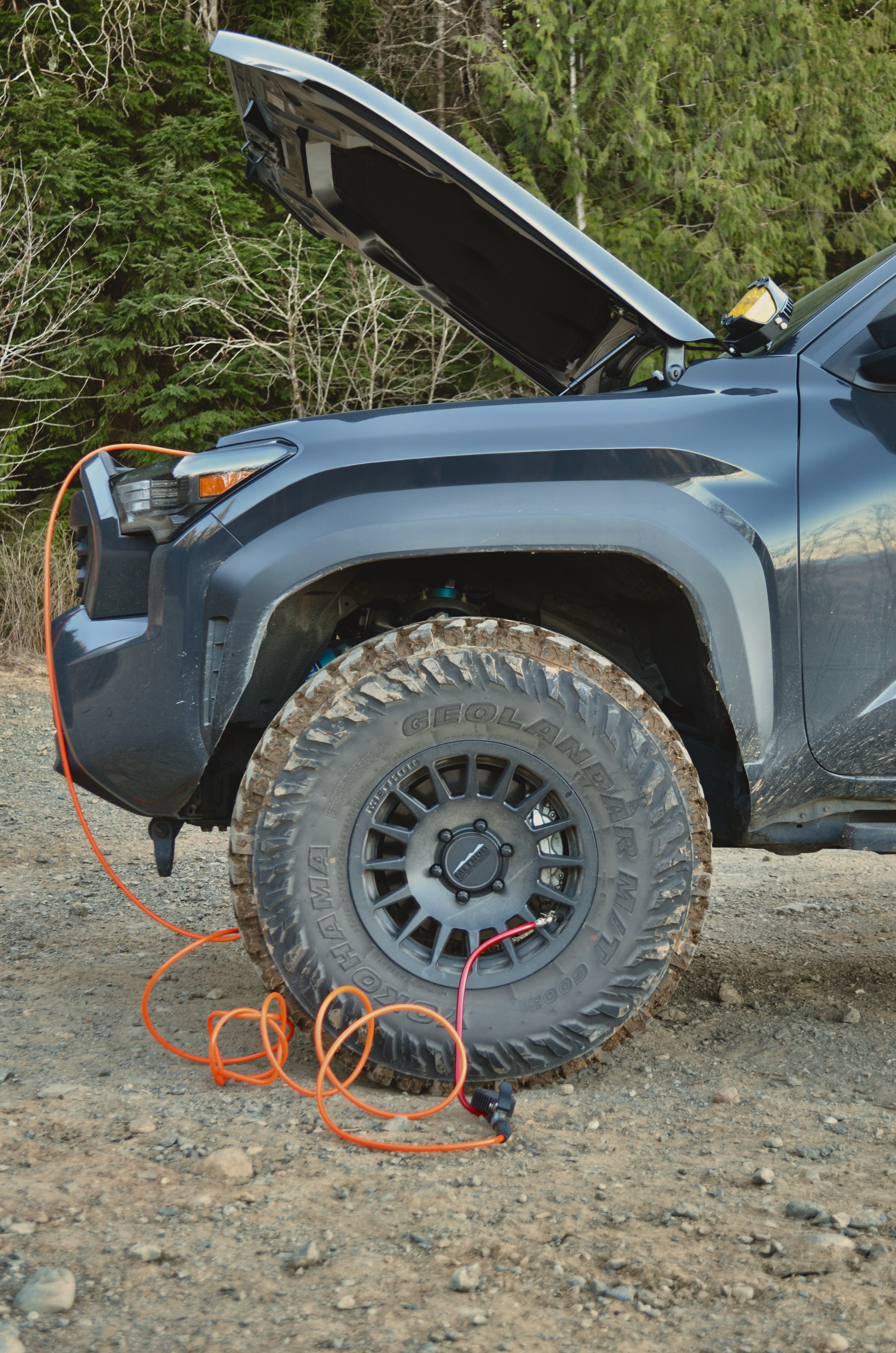 Side view of a blue off-road vehicle with its hood open and an air compressor hose connected to the front left tire, parked on a gravel surface in a wooded area.