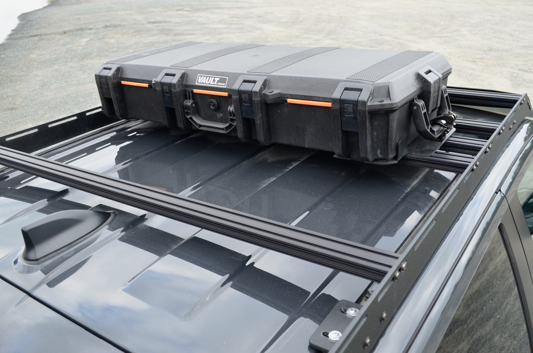 BPF Universal Pelican Vault V700 Case Mounting brackets securing a black protective Pelican Vault case to a roof rack on a dark-colored vehicle, showcasing durable and secure overlanding gear.