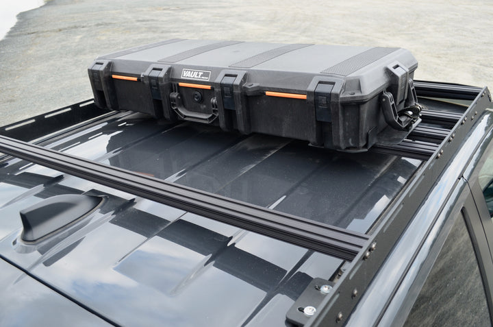 BPF Universal Pelican Vault V700 Case Mounting brackets securing a black protective Pelican Vault case to a roof rack on a dark-colored vehicle, showcasing durable and secure overlanding gear.