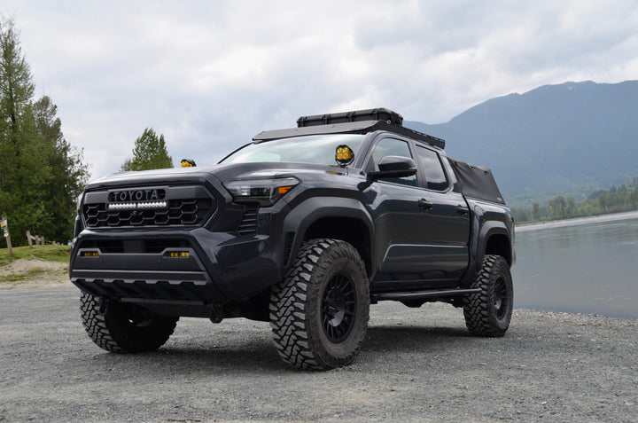 Black Toyota Tacoma truck equipped with off-road tires, roof rack, and accessories, parked by a lakeside with mountains and cloudy sky in the background, showcasing BPF Universal Pelican Vault V700 Case Mounting brackets.