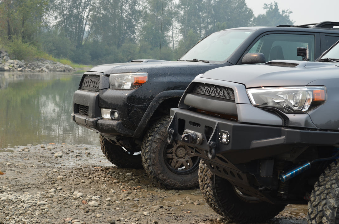 Two Toyota 4Runner SUVs parked on a rocky riverbank with aftermarket bumpers and off-road tires, surrounded by trees and calm water in the background.