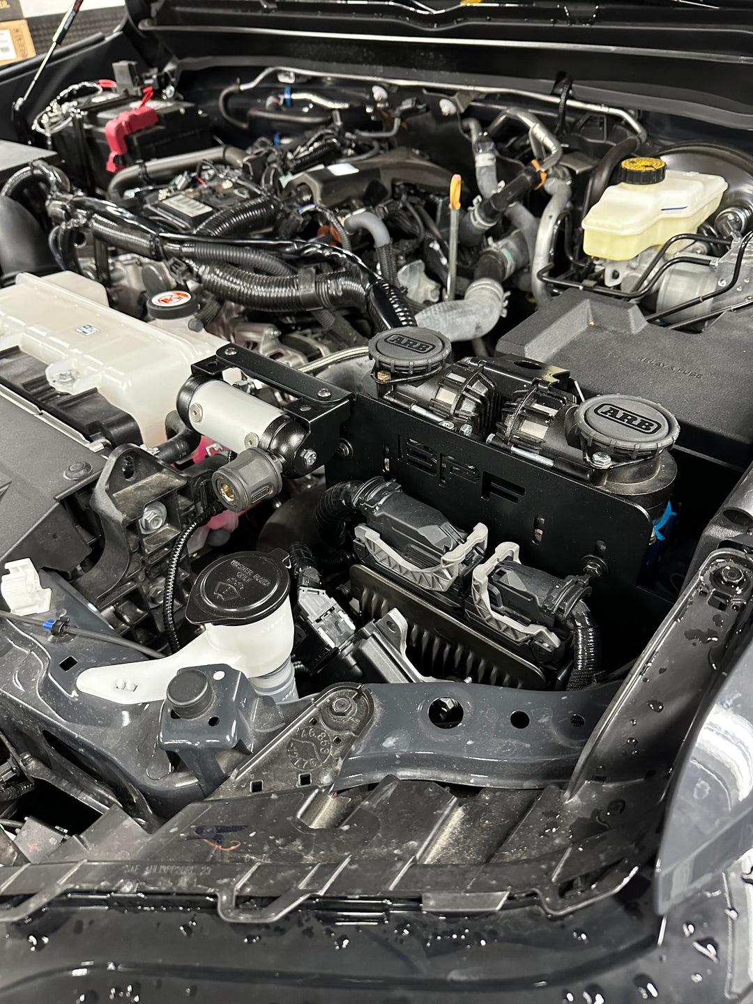 Engine bay featuring a installed BPF 2024-2025 Tacoma and Land Cruiser Twin Compressor Mount with dual compressors secured on a black powder-coated bracket inside a truck.