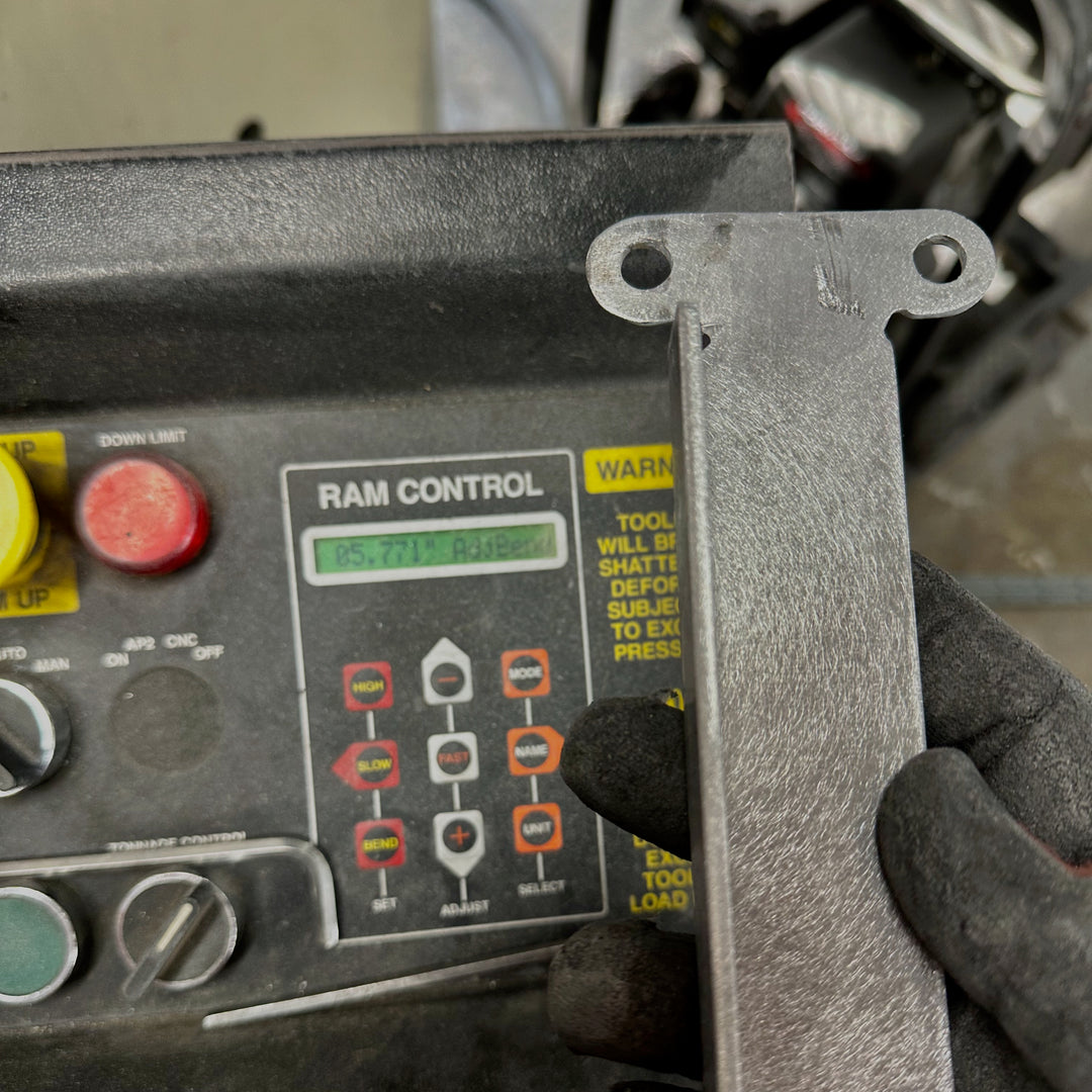 Close-up of a gloved hand holding a flat metal bracket with mounting holes near a machine control panel labeled RAM CONTROL with buttons and a warning label.