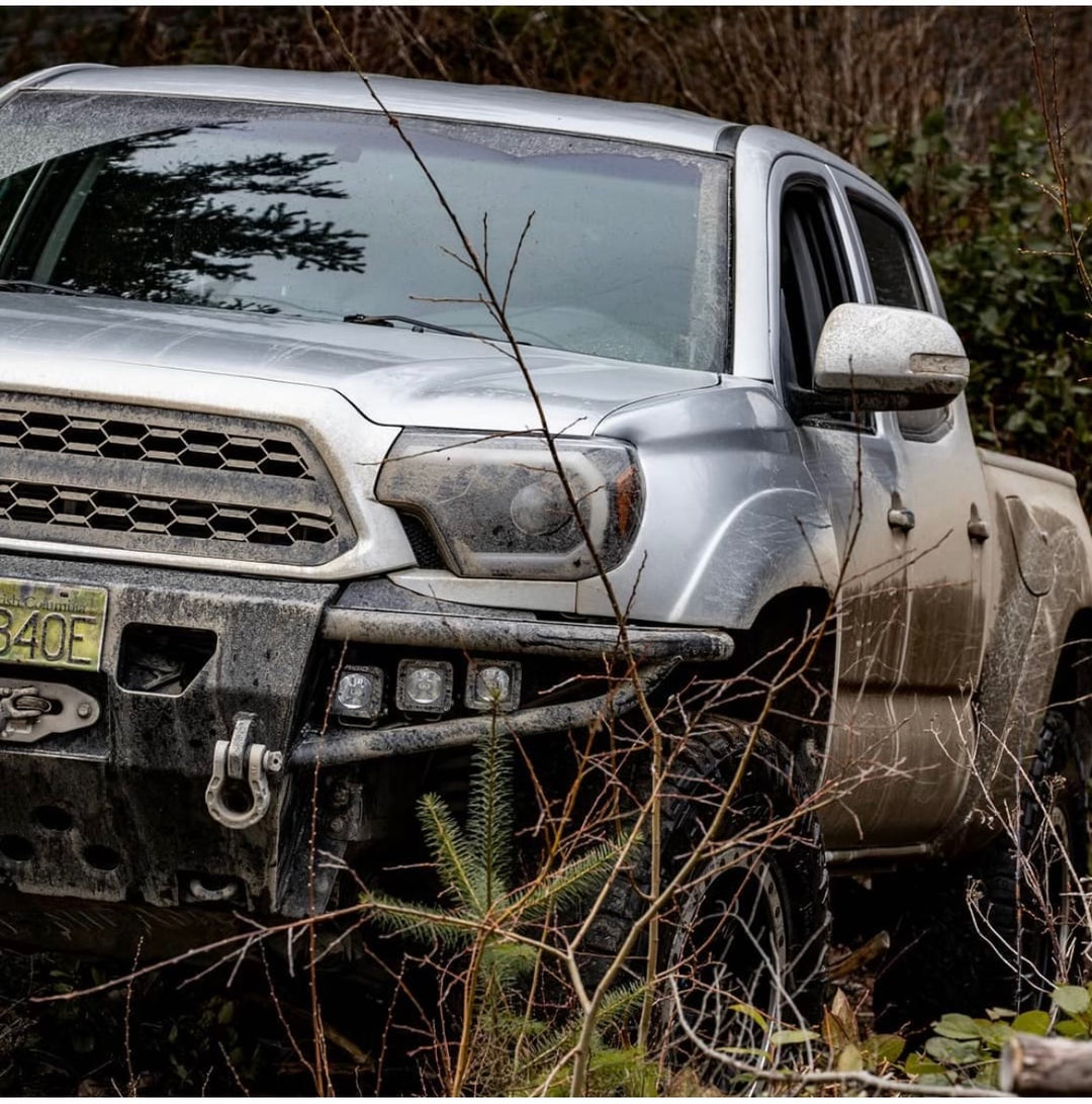 Silver off-road truck covered in mud parked in a wooded area with visible tow hooks and rugged front bumper in natural outdoor setting.