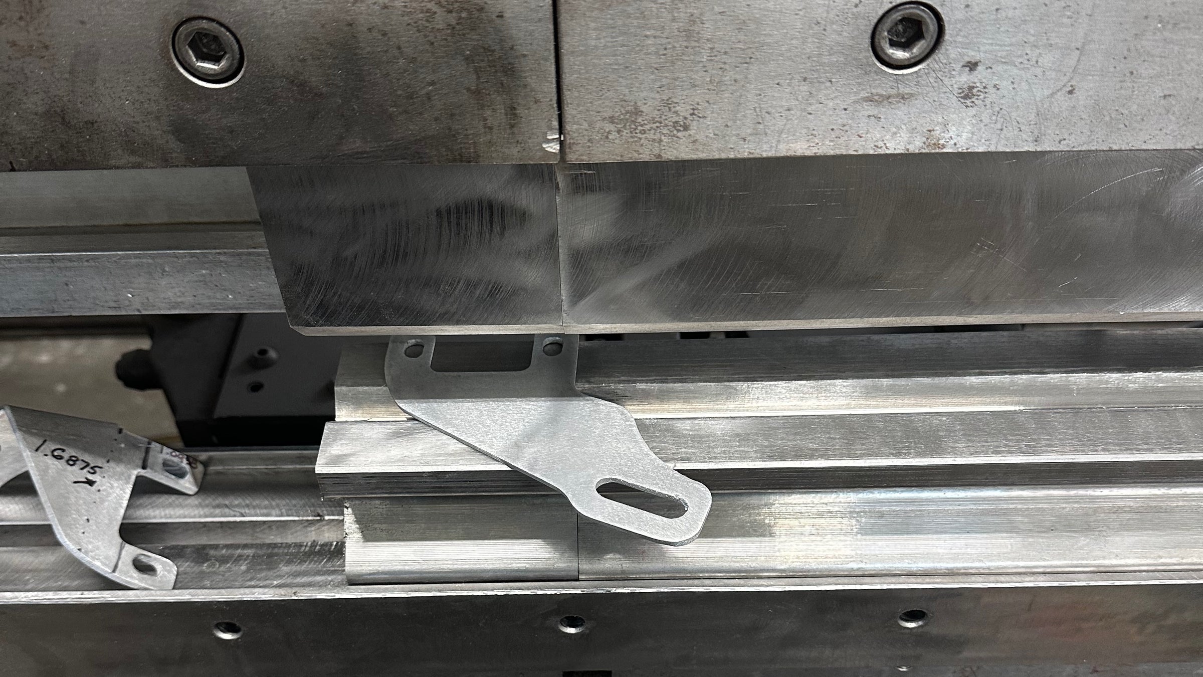Close-up of a metal bracket positioned on an industrial press brake machine, with visible tooling and aluminum extrusion components.
