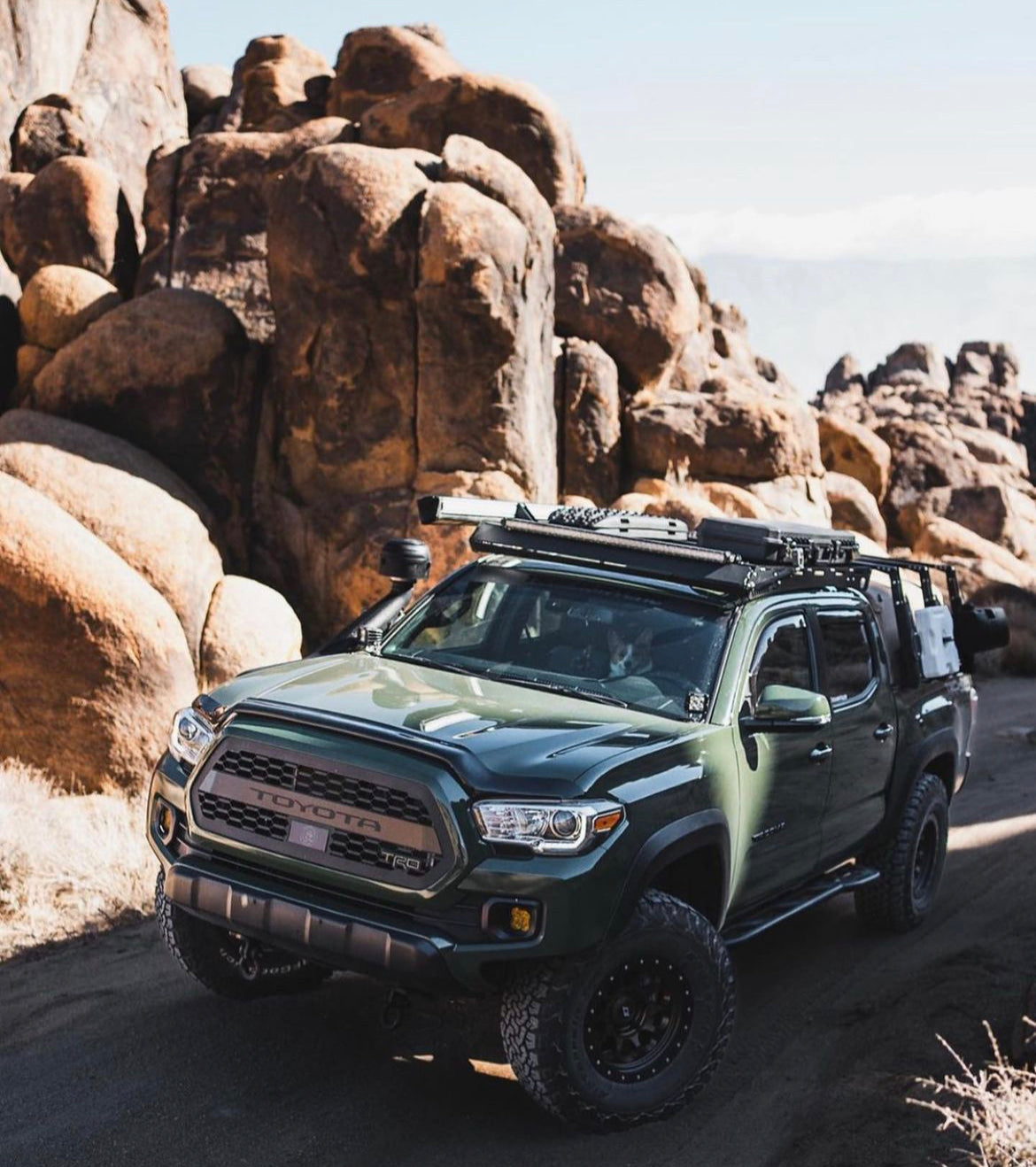 Dark green Toyota Tacoma pickup truck equipped with off-road accessories including roof rack, light bar, and mounted gear, parked on a rocky dirt trail surrounded by large rock formations.