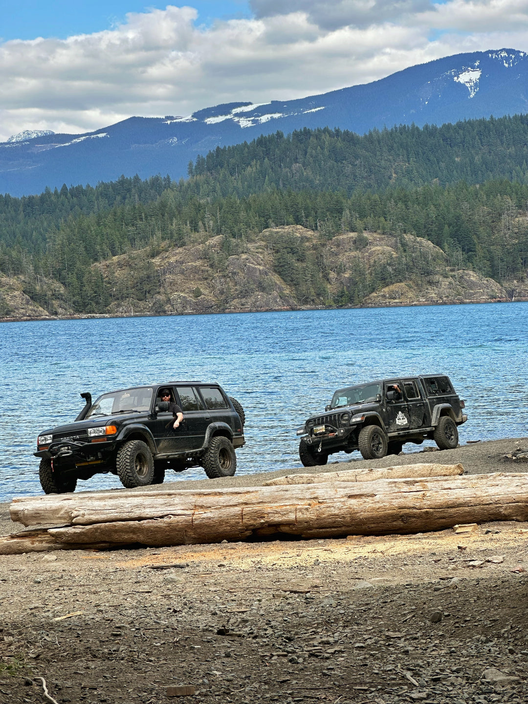 Two black off-road SUVs parked on a rocky lakeshore with a large log in the foreground and a forested mountain landscape with snow-capped peaks in the background under a cloudy sky.