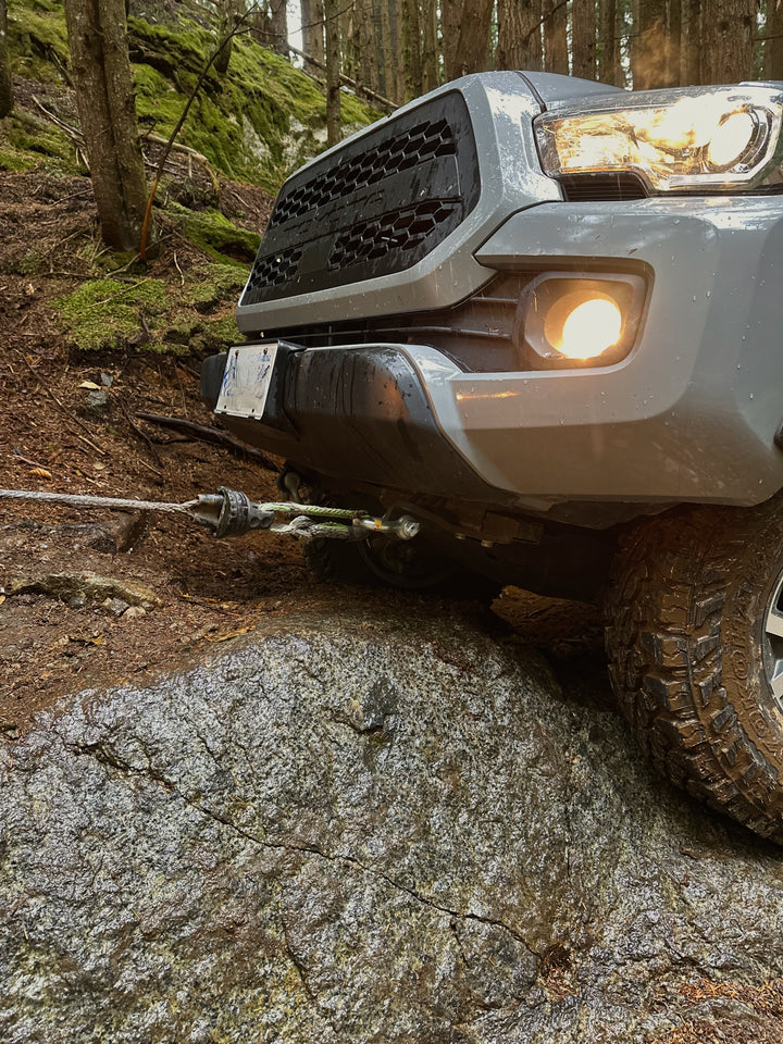 Front view of a silver off-road vehicle with a lit fog light and aggressive tire gripping a large rock, connected to a winch cable, parked on a forest trail with mossy trees.