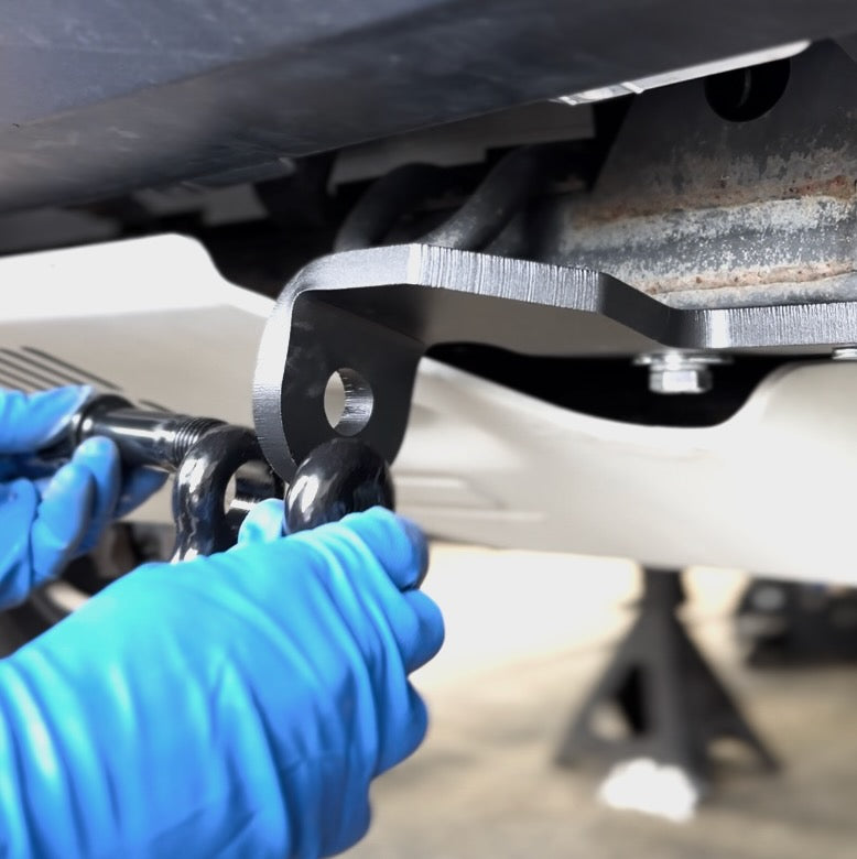 Hands wearing blue gloves installing a black metal tow hook onto a vehicle's rear hitch mount with a visible rusted frame underneath.