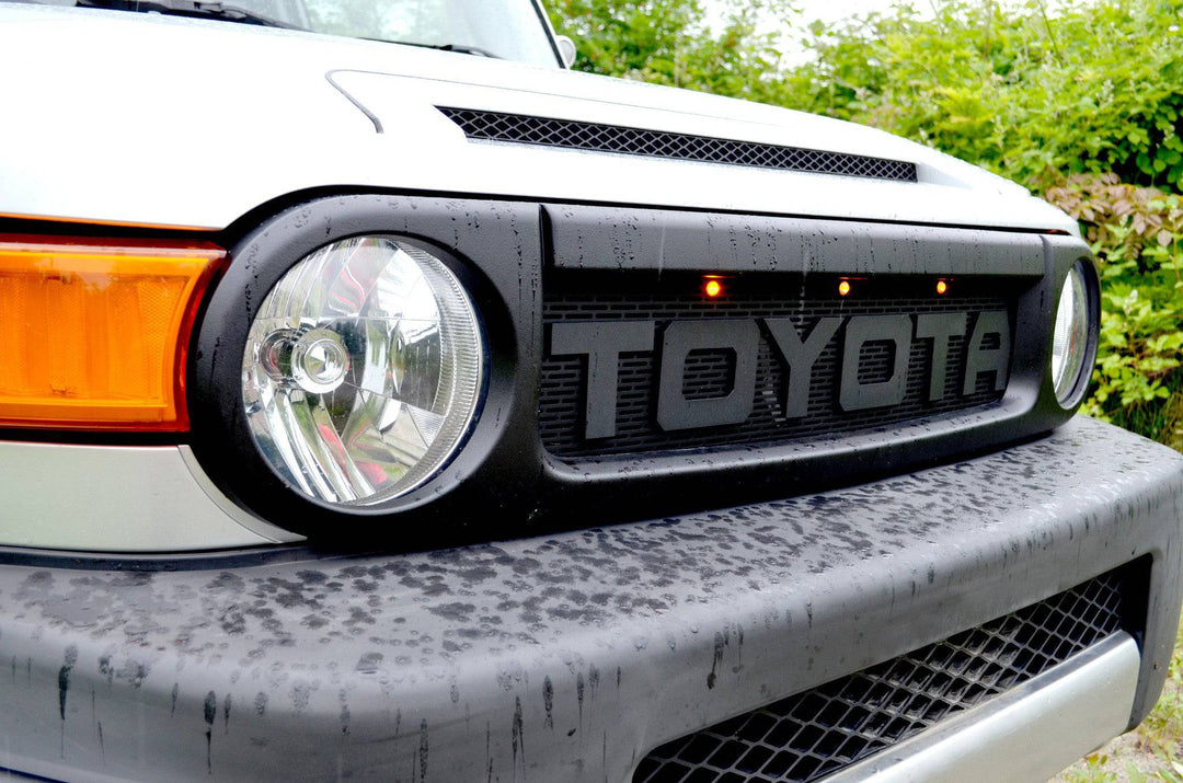 BPF Completed 2007-2014 FJ Cruiser Grill installed on front of white Toyota FJ Cruiser, featuring rugged black finish and bold TOYOTA lettering with three amber marker lights above.