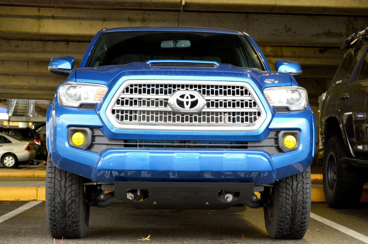 Blue Toyota Tacoma truck front view showing installed BPF 2016-2023 Tacoma Skid Plate under the bumper in a parking garage with powder-coated finish and OEM fitment.