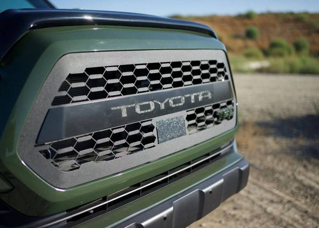 Close-up of a black and matte gray BPF 2016-2023 Toyota Tacoma Grill with hexagonal mesh pattern and TOYOTA lettering, installed on an olive green truck outdoors.