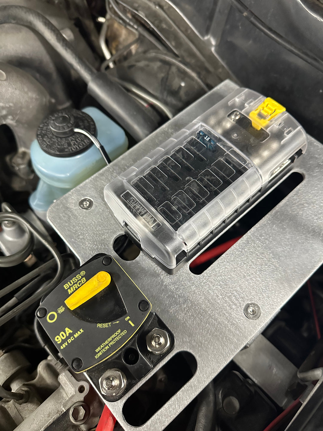 Close-up view of a BPF 80 series Land cruiser Fuse block mount installed in a vehicle engine bay, featuring a clear plastic fuse holder and a yellow resettable 90A circuit breaker for electrical protection.