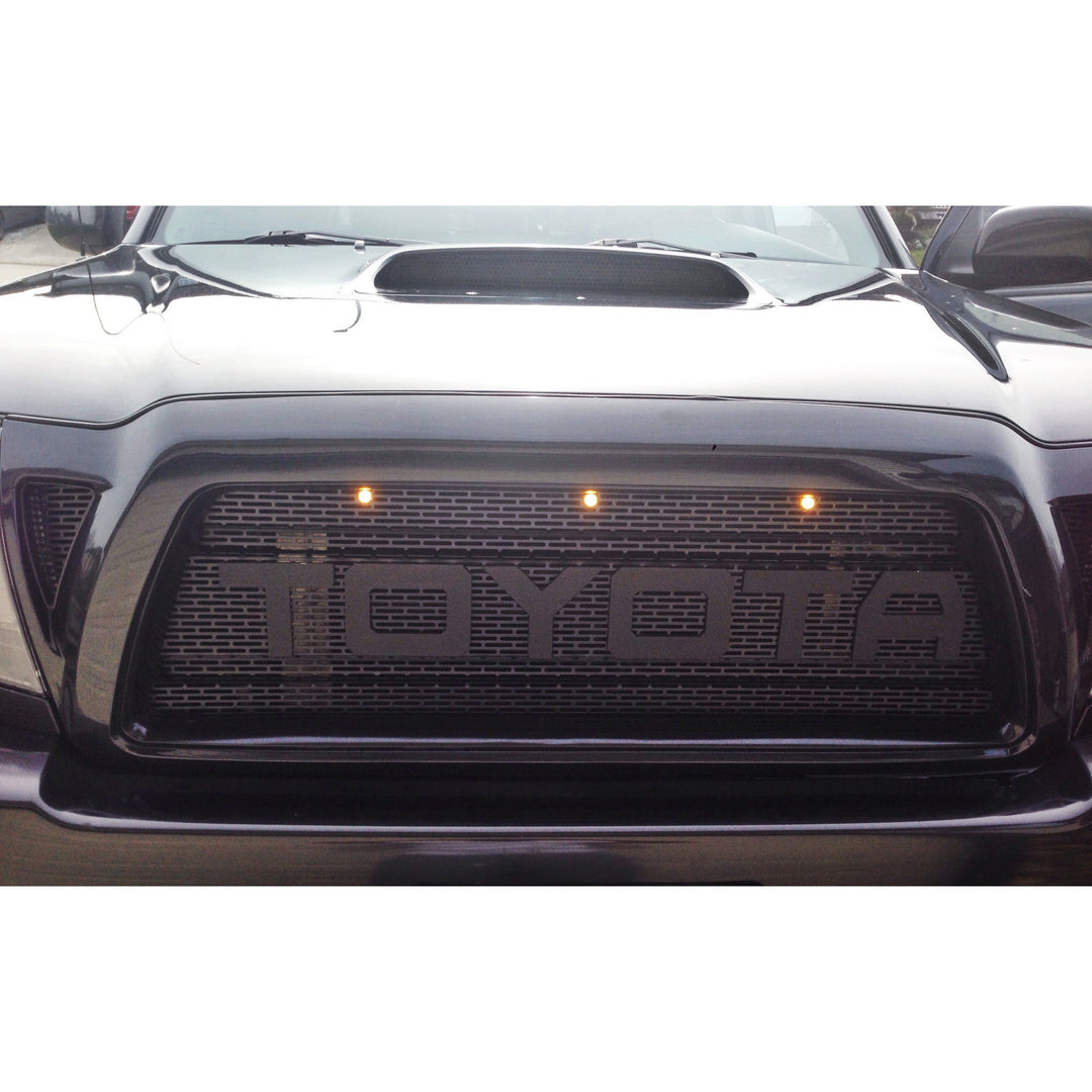 Close-up of a black truck grille featuring the BPF 2005-2011 Toyota Tacoma Raptor Style Mesh and Lettering with integrated lights and a rugged, aggressive design.