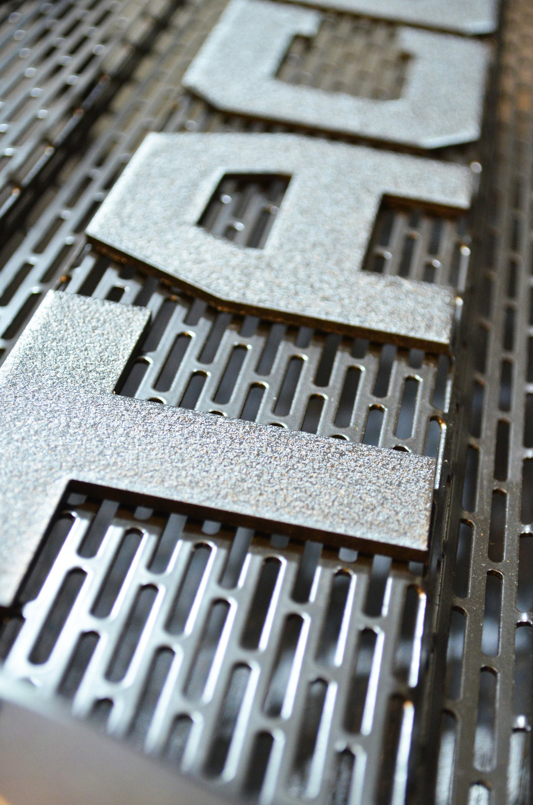 Close-up of textured silver lettering mounted on a black raptor style grill mesh, showcasing the design detail of the BPF 2005-2011 Toyota Tacoma Raptor Style Mesh and Lettering accessory.