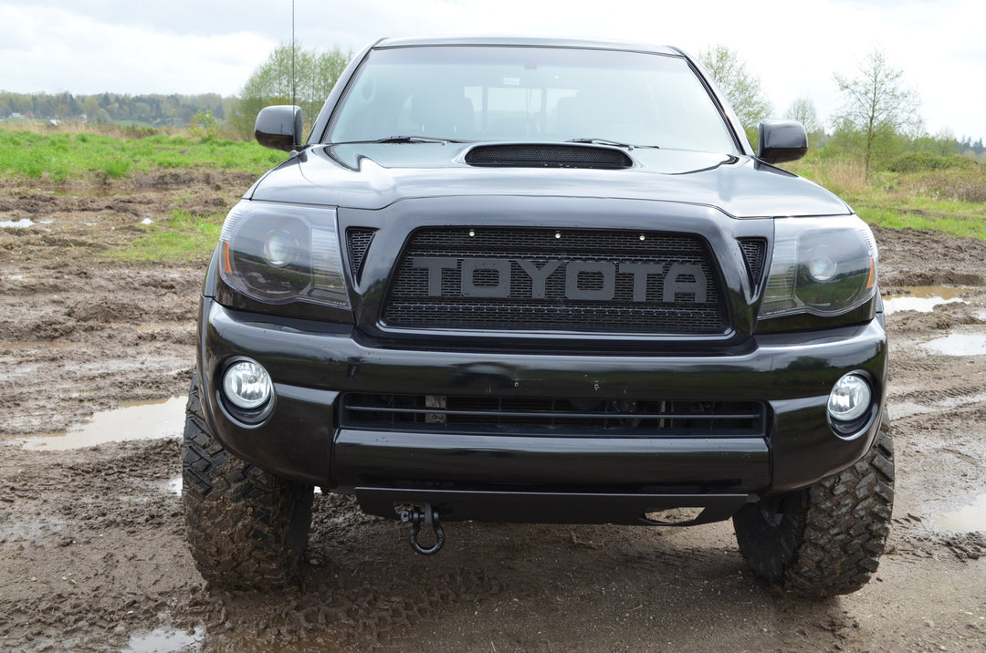 Front view of a black Toyota Tacoma equipped with BPF 2005-2011 Toyota Tacoma Raptor Style Mesh and Lettering grill in a muddy outdoor setting.