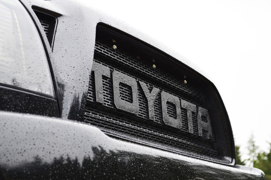 Close-up view of a black truck front grille with large bold lettering spelling TOYOTA, featuring the BPF 2005-2011 Toyota Tacoma Raptor Style Mesh and Lettering in a textured black finish with water droplets on the surface.