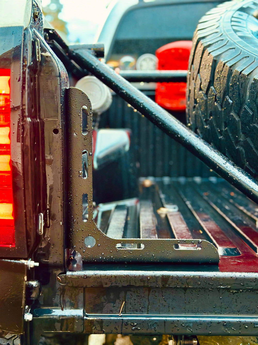 Close-up of a wet truck bed showing the BPF 2005-2023 Toyota Tacoma Bed Stiffeners installed near the tailgate with off-road gear and spare tire in the background.