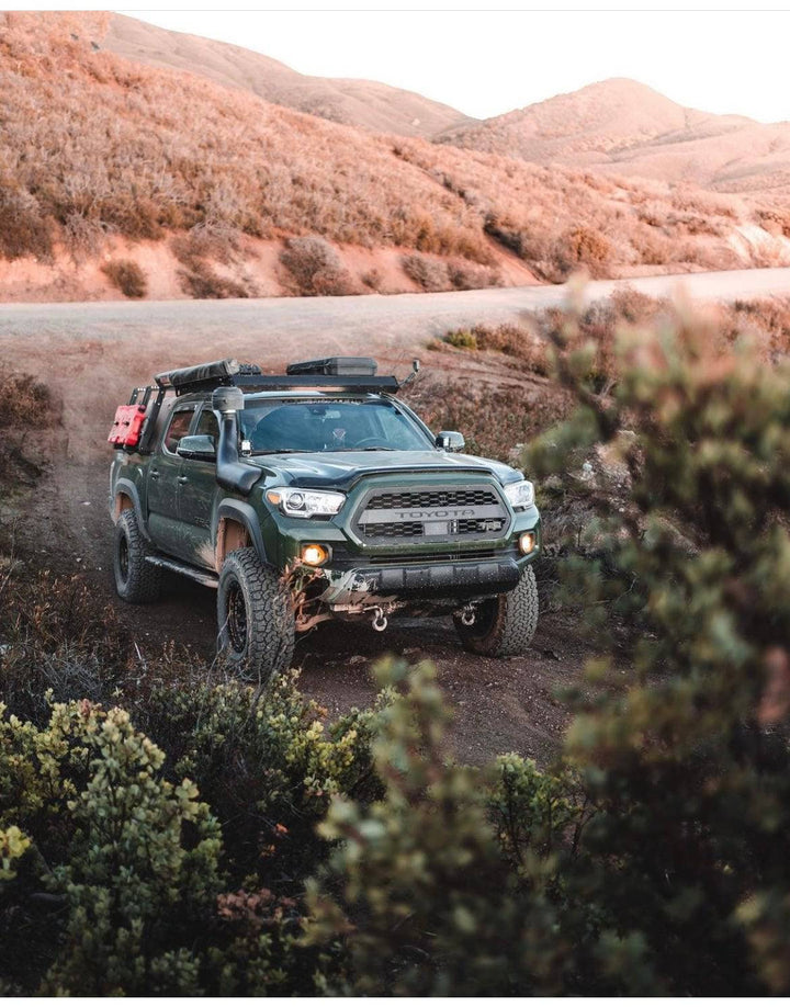 Off-road Toyota Tacoma featuring a rugged front with BPF 2016-2023 Toyota Tacoma Grill, equipped for adventure with off-road tires, roof rack gear, and front tow hooks.